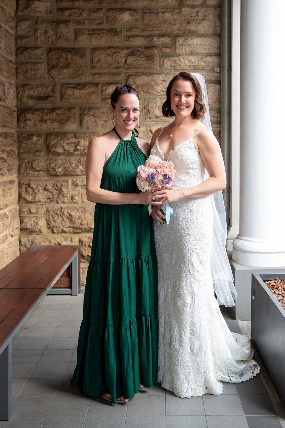 Bride in white lace gown holds pink bouquet beside bridesmaid in emerald green halter dress against stone wall.