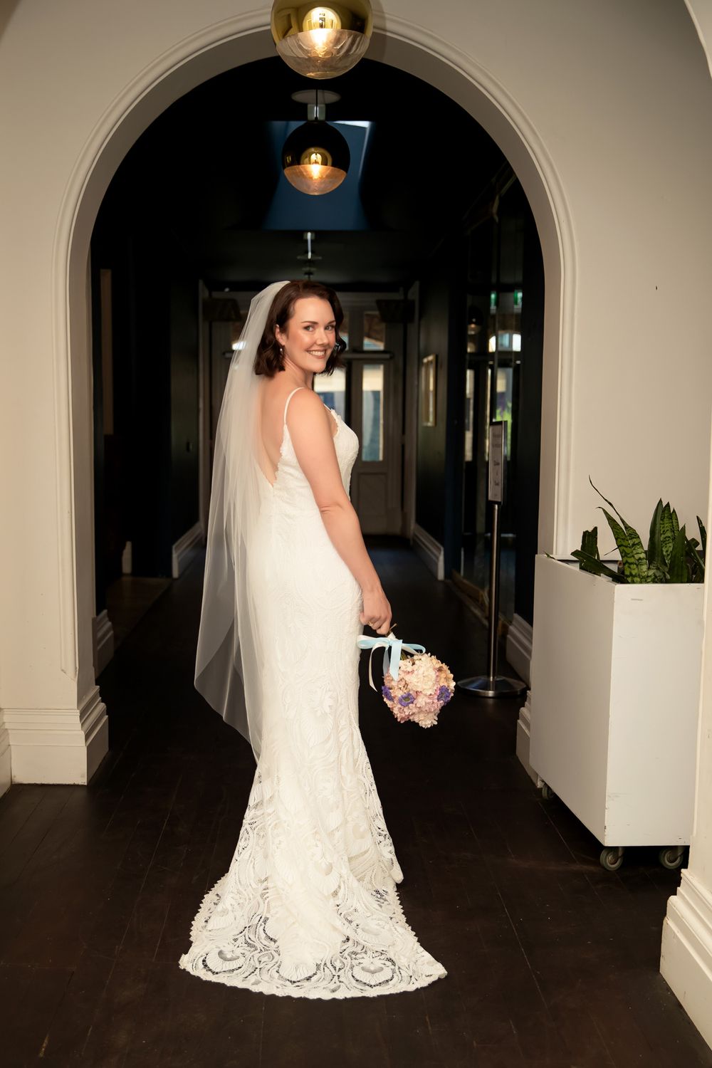 Bride in white lace gown and veil holds bouquet, posing under elegant arch in a venue hallway.