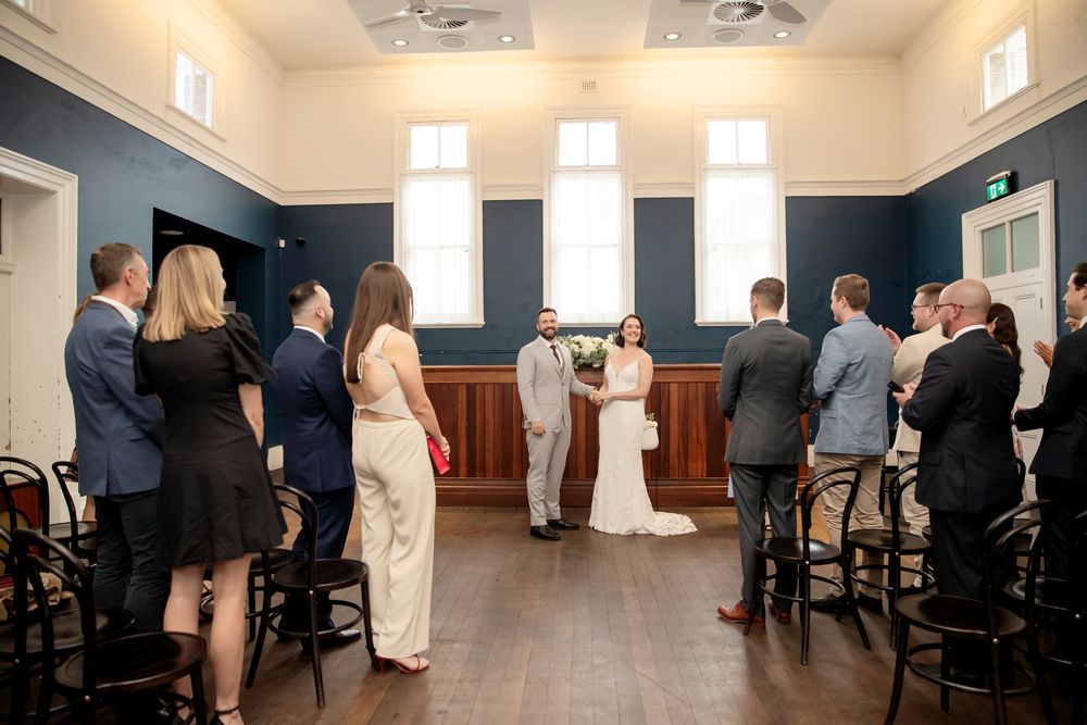 Intimate wedding ceremony in a elegant blue room with wooden floors, guests standing as couple exchanges vows.