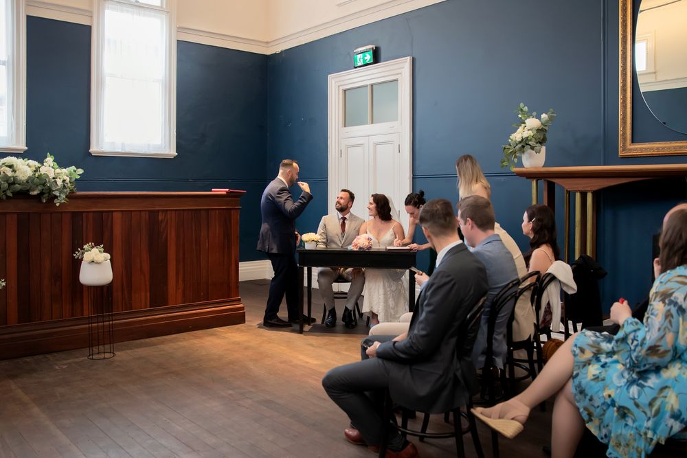 Intimate civil wedding ceremony in a blue-walled room with guests seated as officiant speaks to couple at table.
