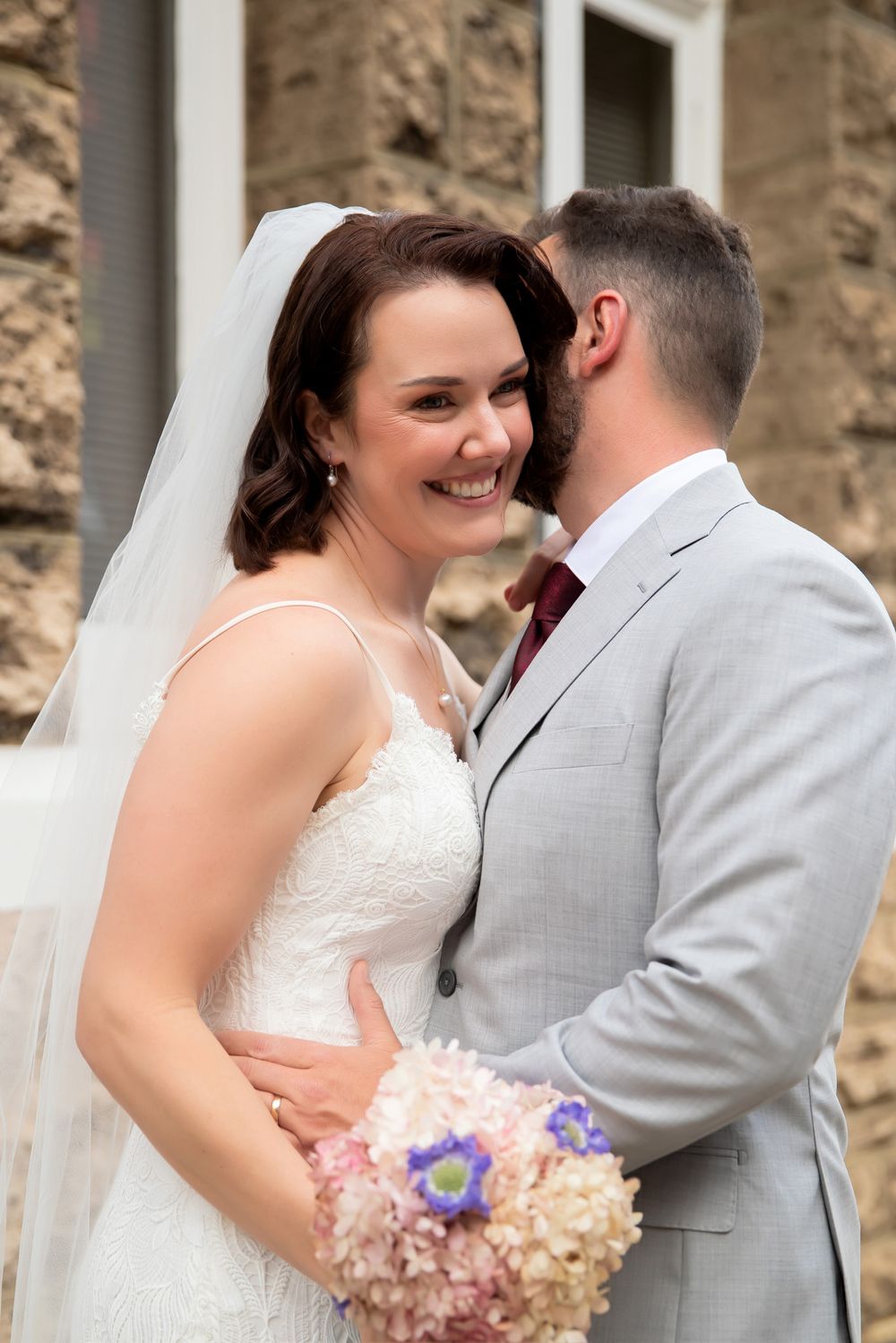 Smiling bride in white gown holding pink bouquet embraced by groom in gray suit with burgundy tie outdoors.