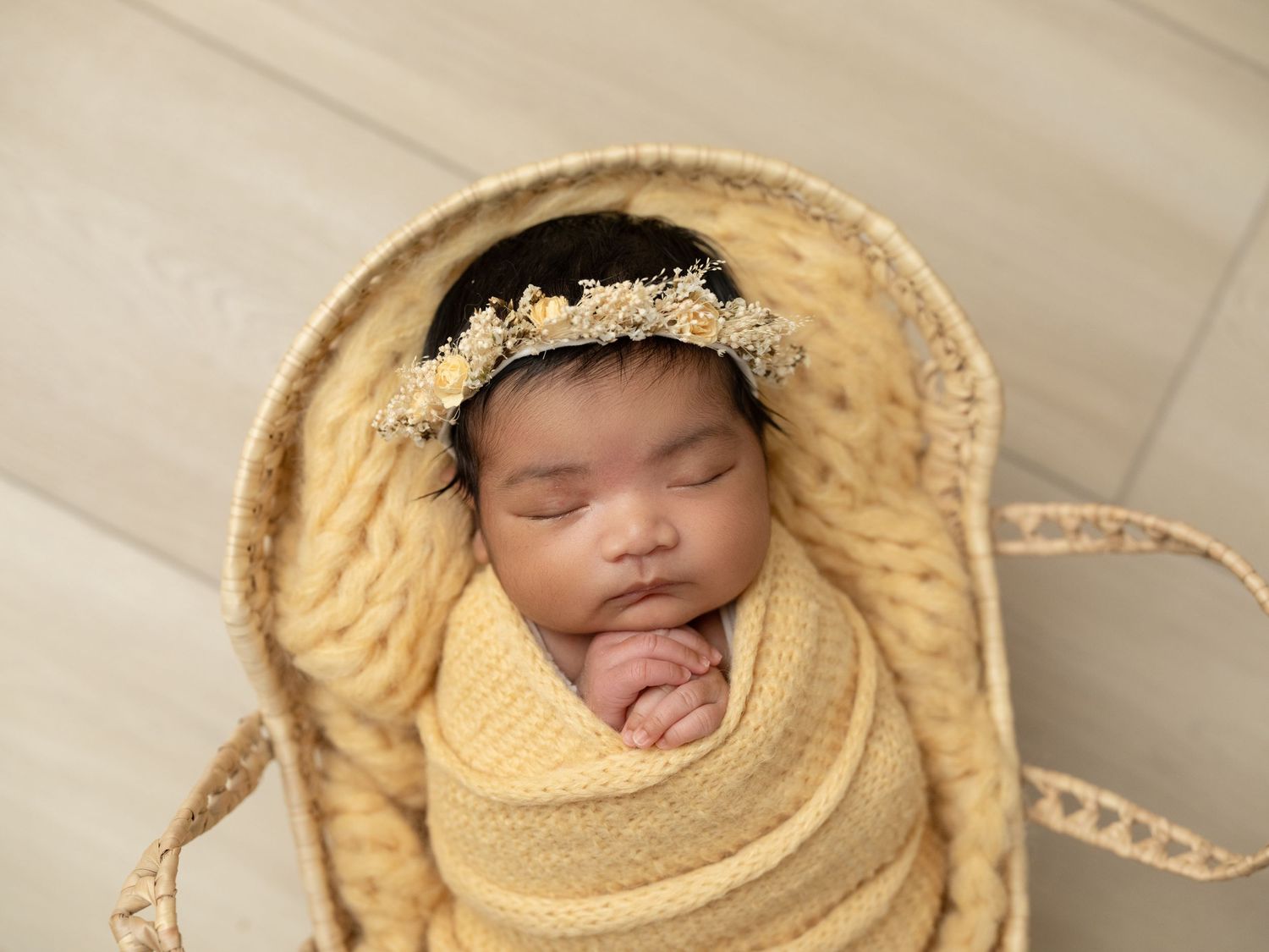 Sleeping newborn baby swaddled in yellow knit wrap with floral headband, nestled in a wicker basket.