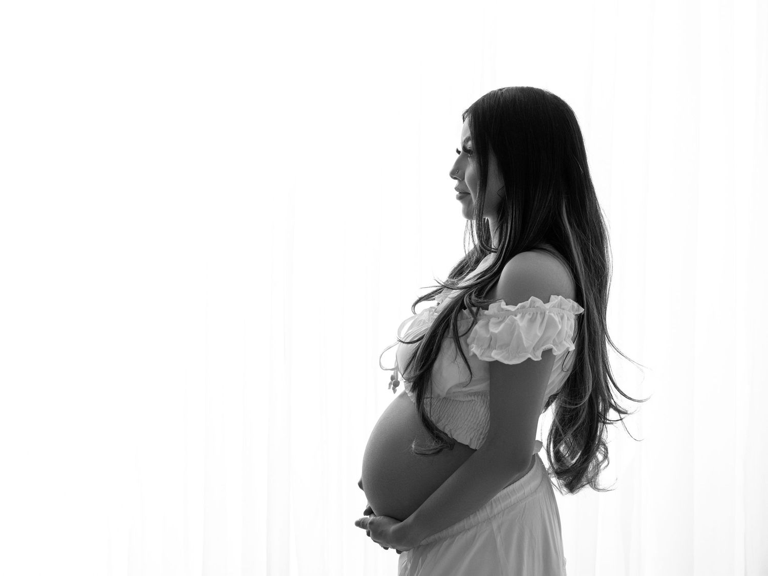 Pregnant woman in white off-shoulder dress cradling baby bump, standing against soft white curtains.