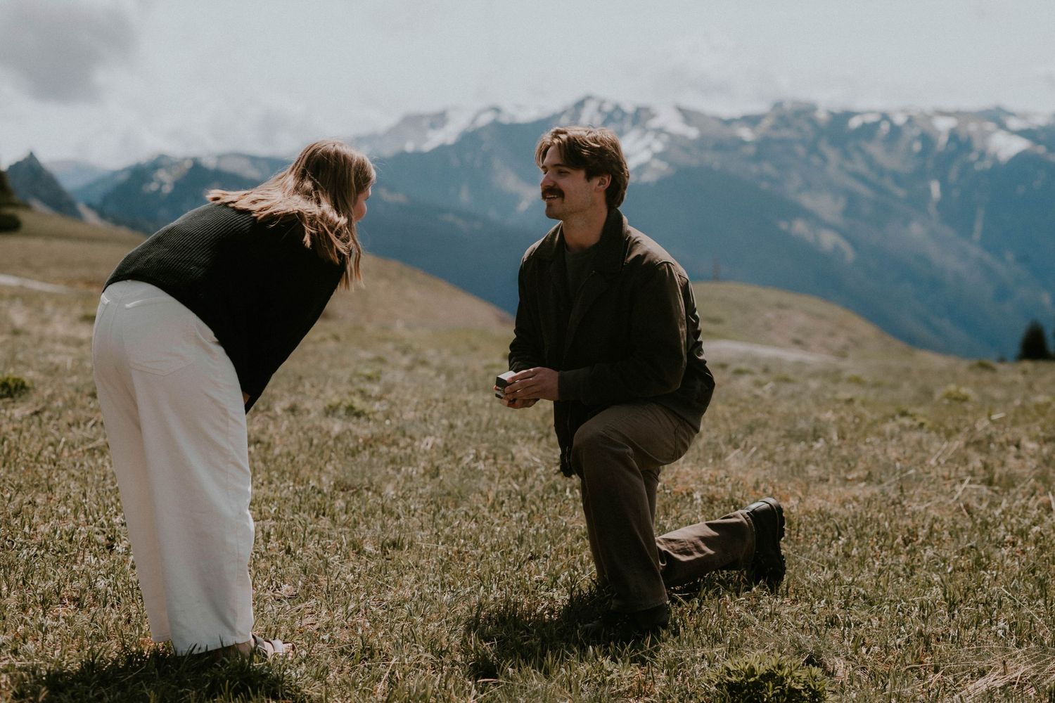 Man kneeling on mountain meadow proposing to woman with snow-capped peaks in background during outdoor engagement.