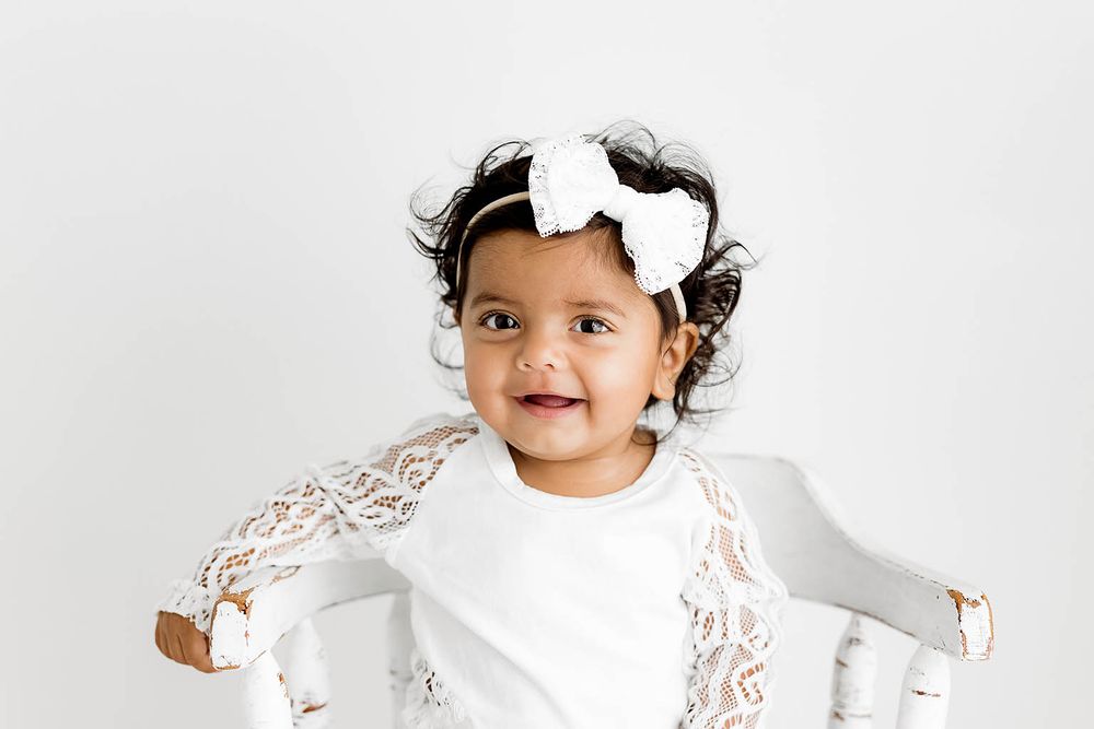 Smiling toddler girl in white lace dress and bow headband sitting on white chair against light background.