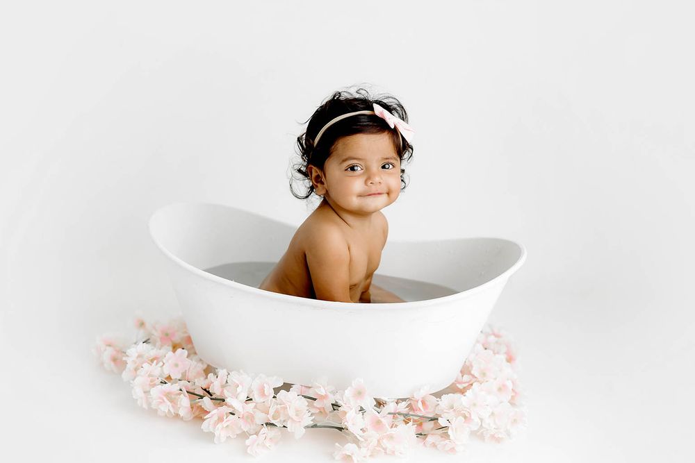 Adorable baby girl with floral headband sitting in white tub with pink blossoms.