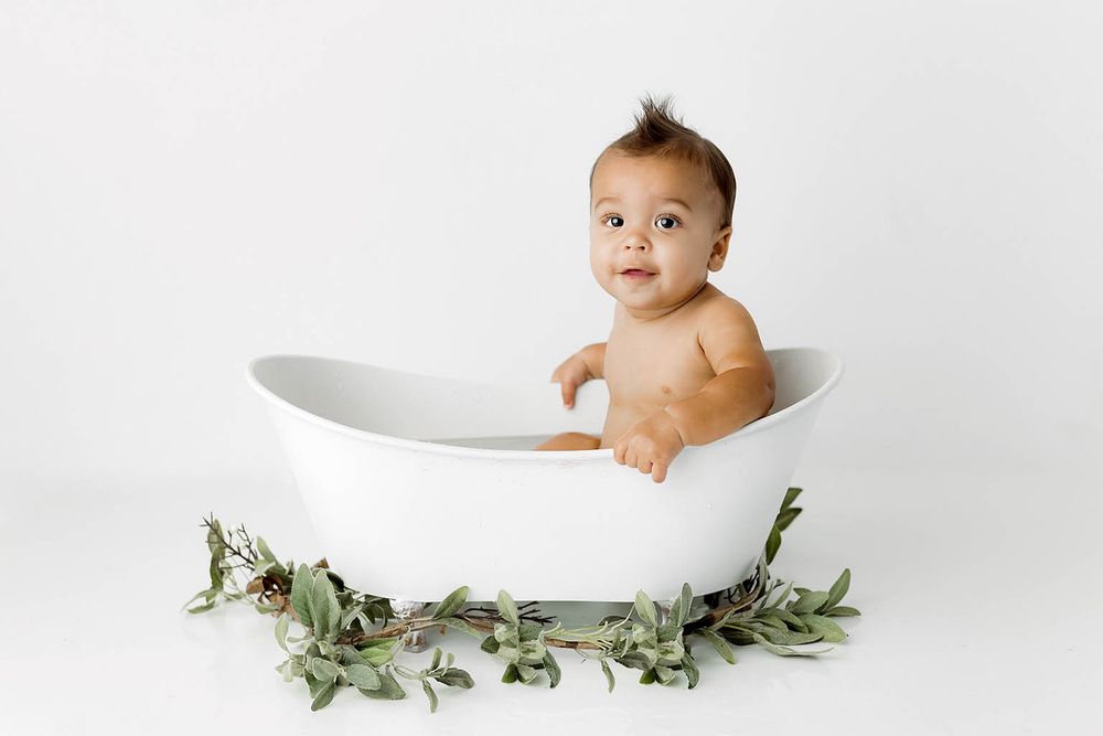 Cute baby sitting in a white bathtub surrounded by green leaves on a white background.