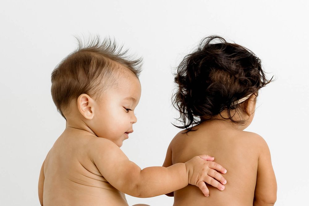 Two babies in diapers sitting together, one touching the other's back, against a white background.
