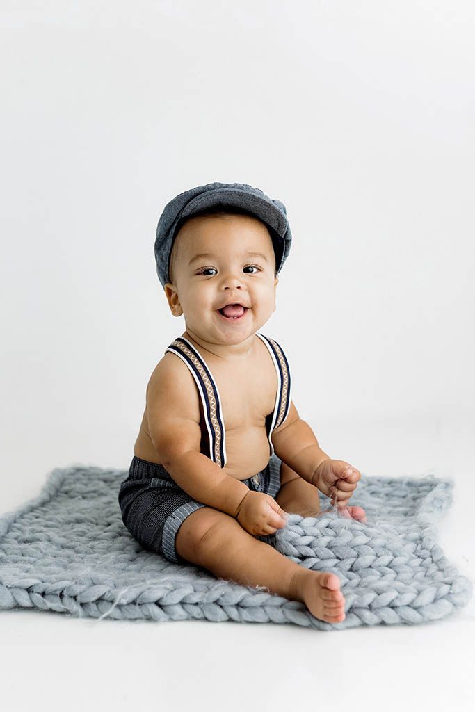 Smiling baby boy in gray shorts, suspenders, and newsboy cap sitting on chunky knit blanket against white background.