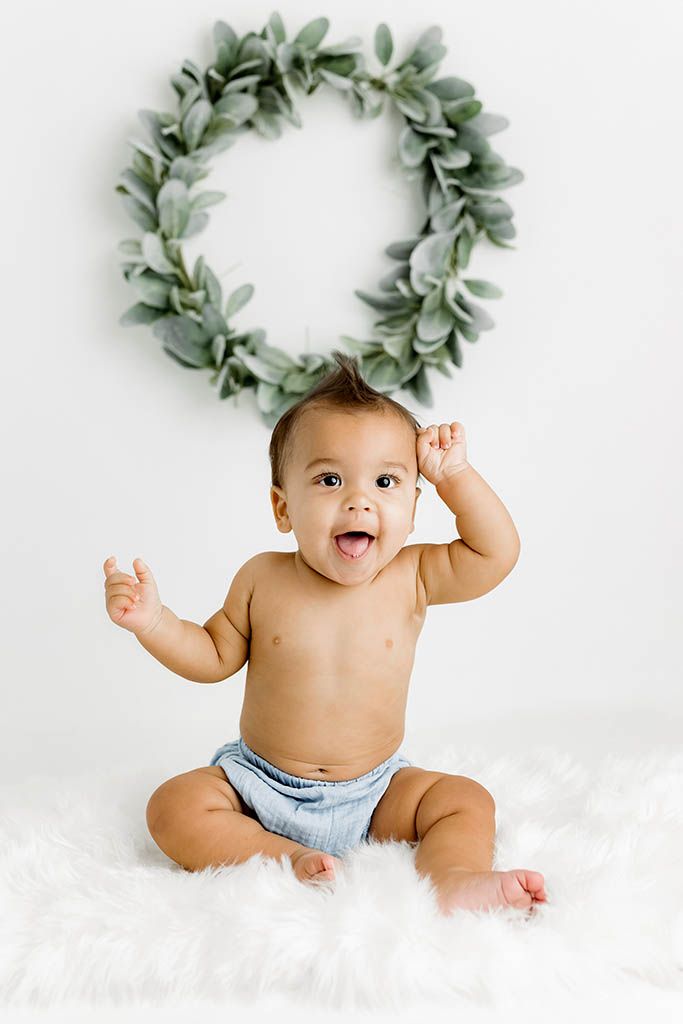 Happy baby sitting on white fur rug in diaper, arms raised, with green wreath backdrop.