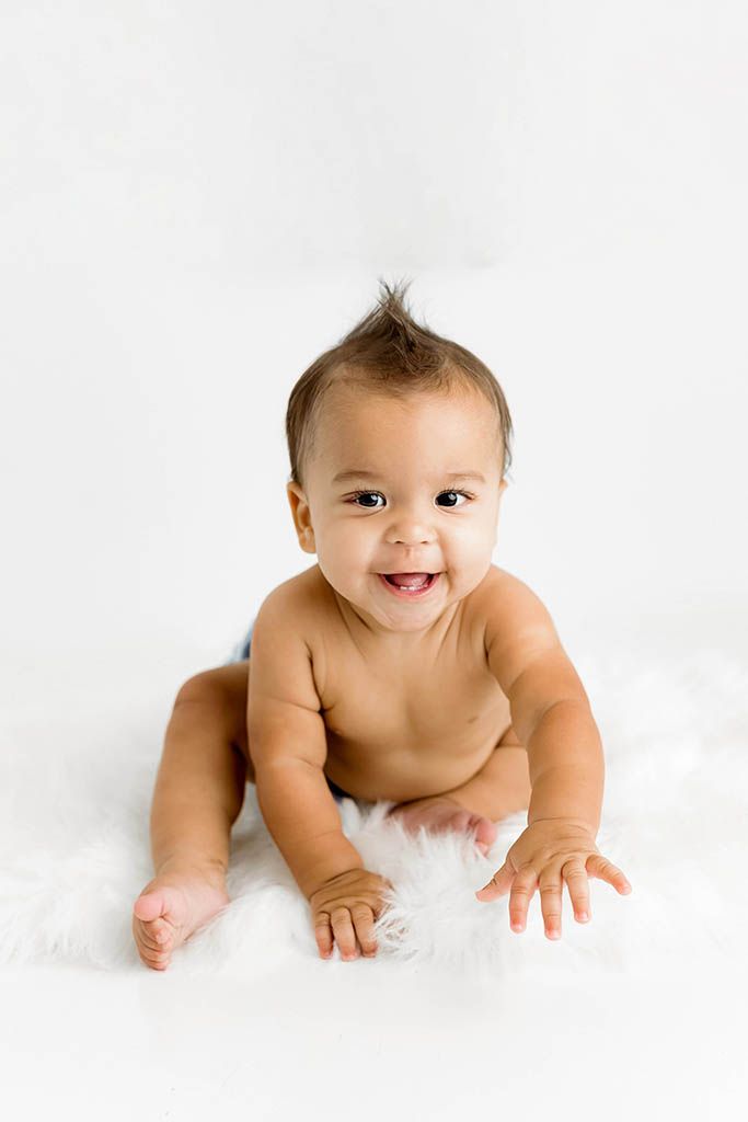 Smiling baby in diaper sitting on white fluffy rug against white background, looking at camera.