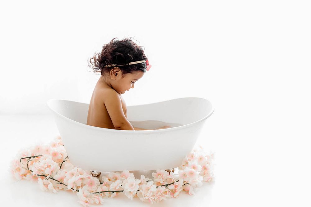 Cute baby girl sitting in a white bathtub surrounded by pink flowers during a milk bath photo session.