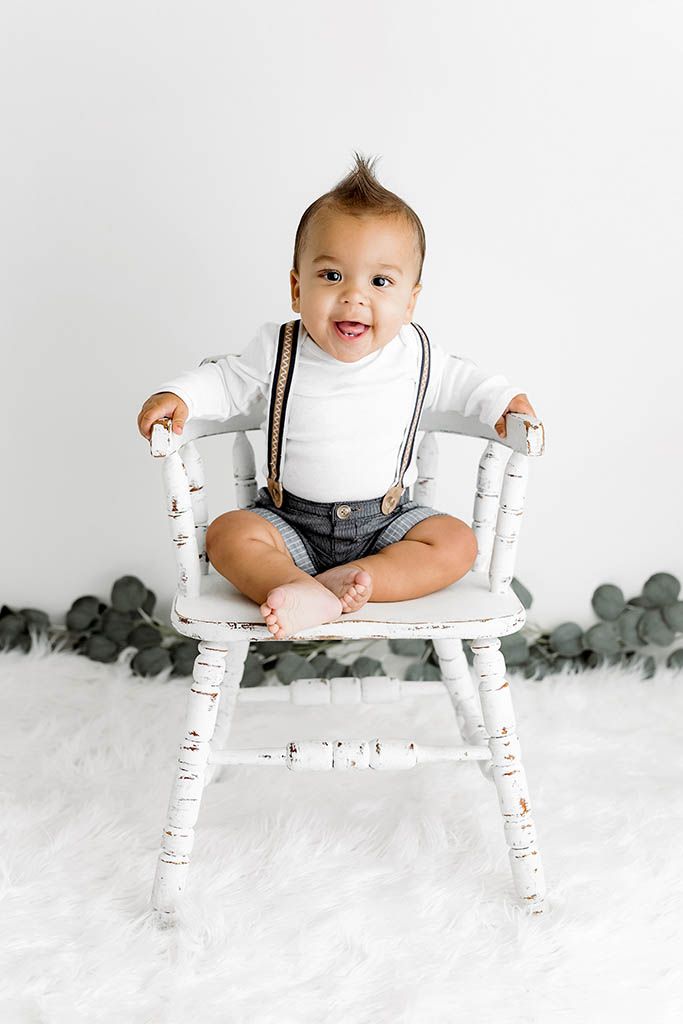 Smiling baby boy in white onesie with suspenders and denim shorts sitting on a rustic white wooden chair.