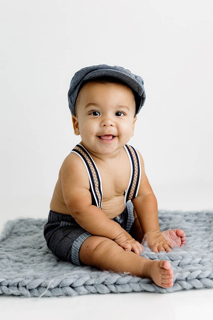 Smiling baby boy in gray newsboy cap and suspenders sits on chunky knit blanket against white background.