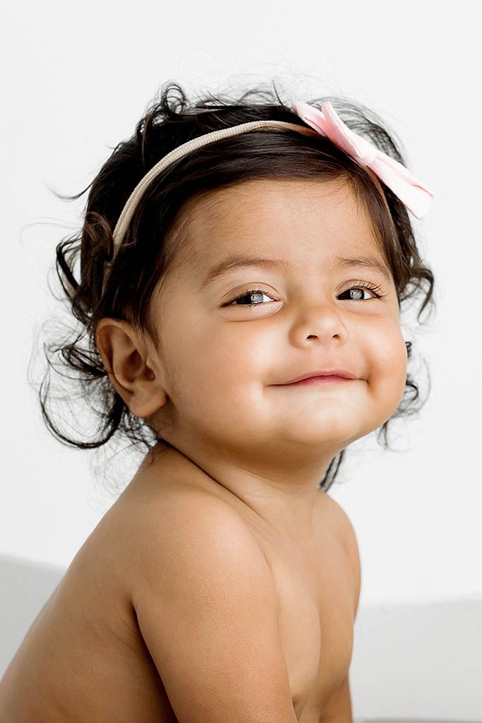 Adorable baby girl with dark curly hair and pink bow headband smiling sweetly against white background.