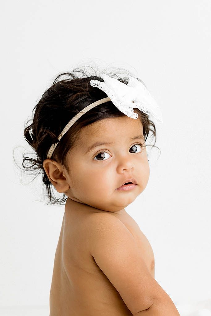 Adorable baby girl with white bow headband and dark curly hair posing against white background.