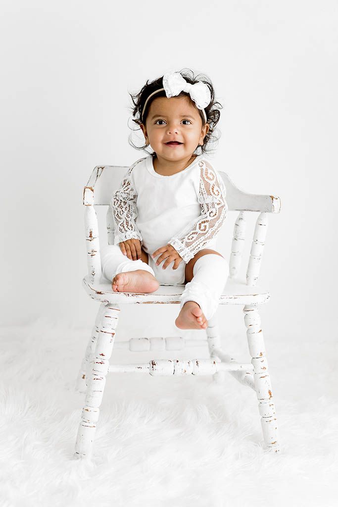 Adorable baby girl in white lace outfit sitting on a rustic white wooden chair with a bow headband.