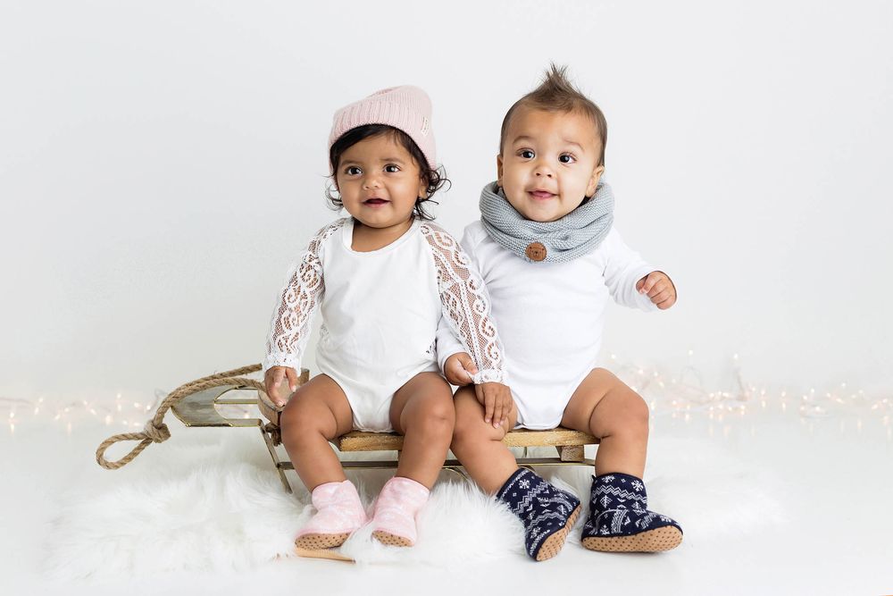 Two adorable babies sitting together on a small sled wearing white outfits with colorful accessories.