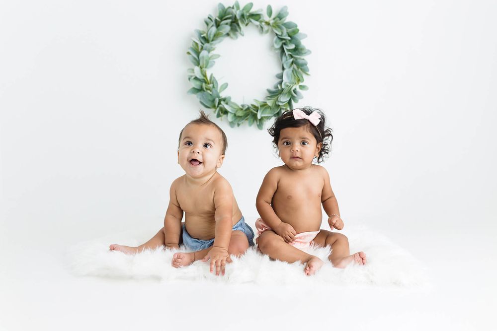 Two adorable babies in diapers sit on white fluff before a green wreath backdrop in a festive holiday photo.
