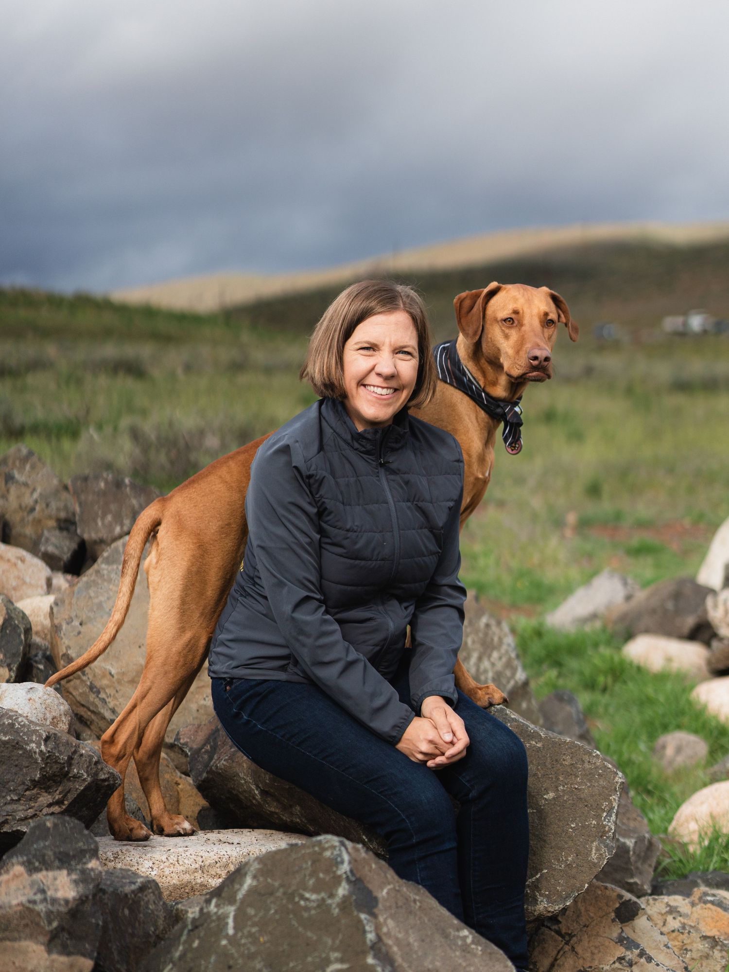 Woman smiling on rocks with a Rhodesian Ridgeback dog leaning on her back in a scenic outdoor landscape.