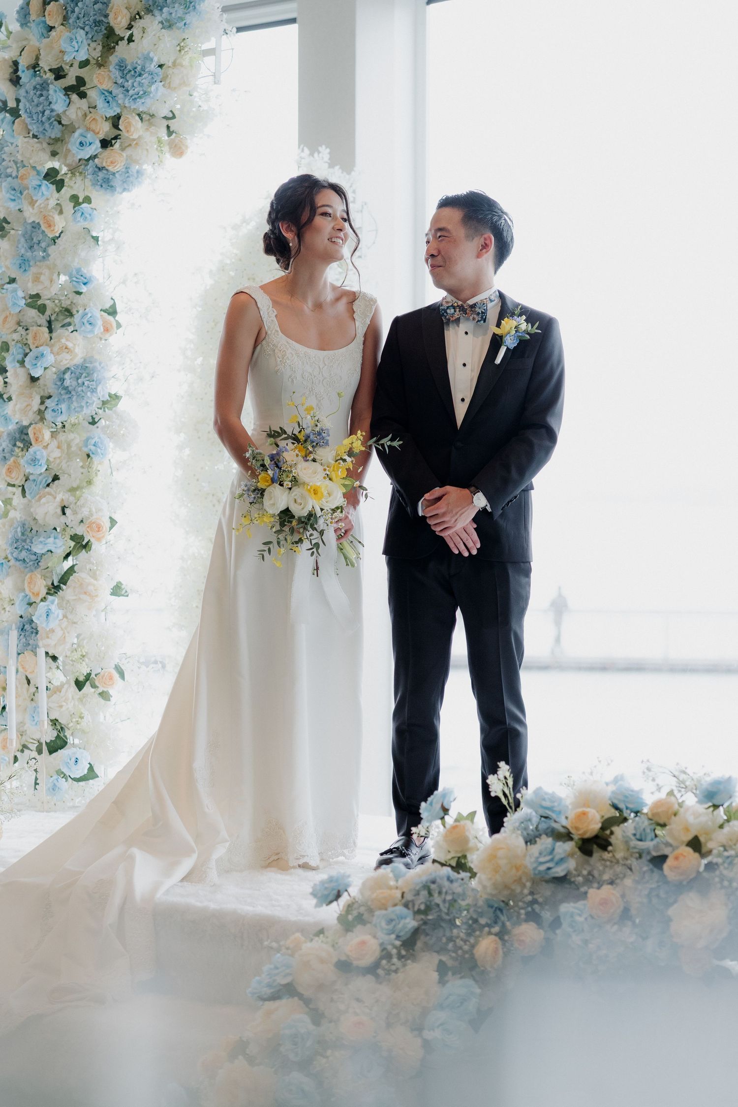 A couple poses playfully under white arched columns in matching light-colored outfits.