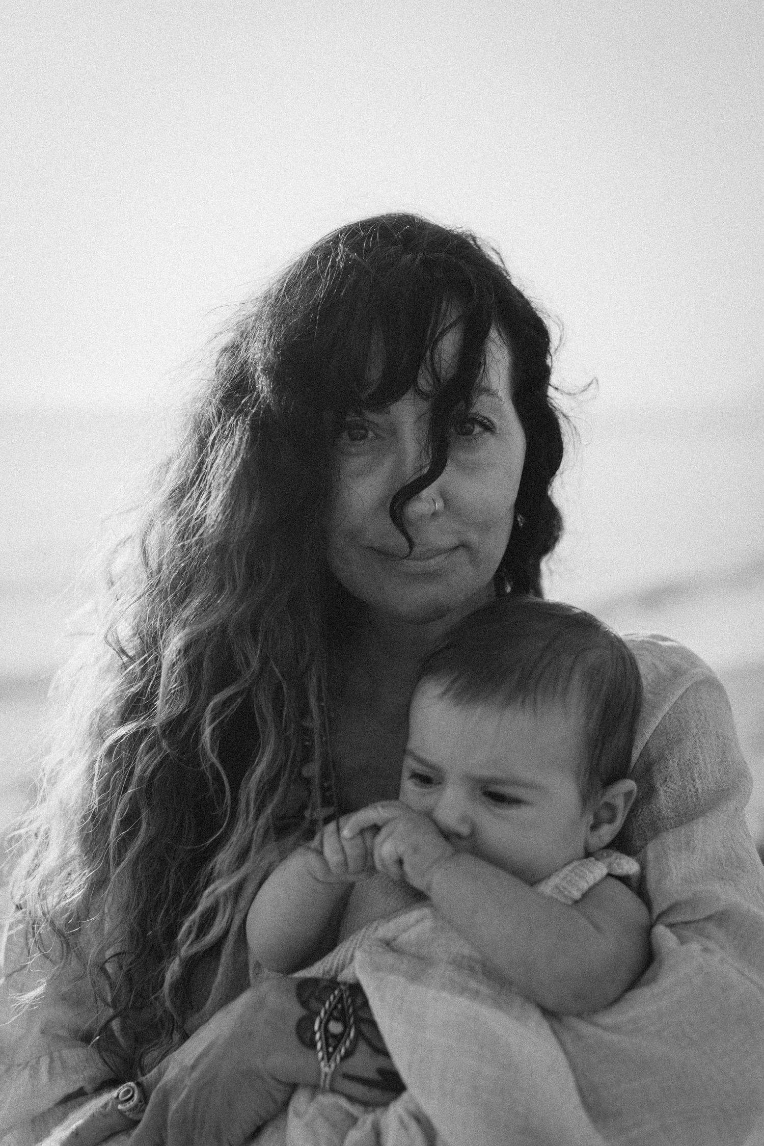 Black and white portrait of a woman with long curly hair tenderly holding a baby wrapped in white cloth Javea photographer