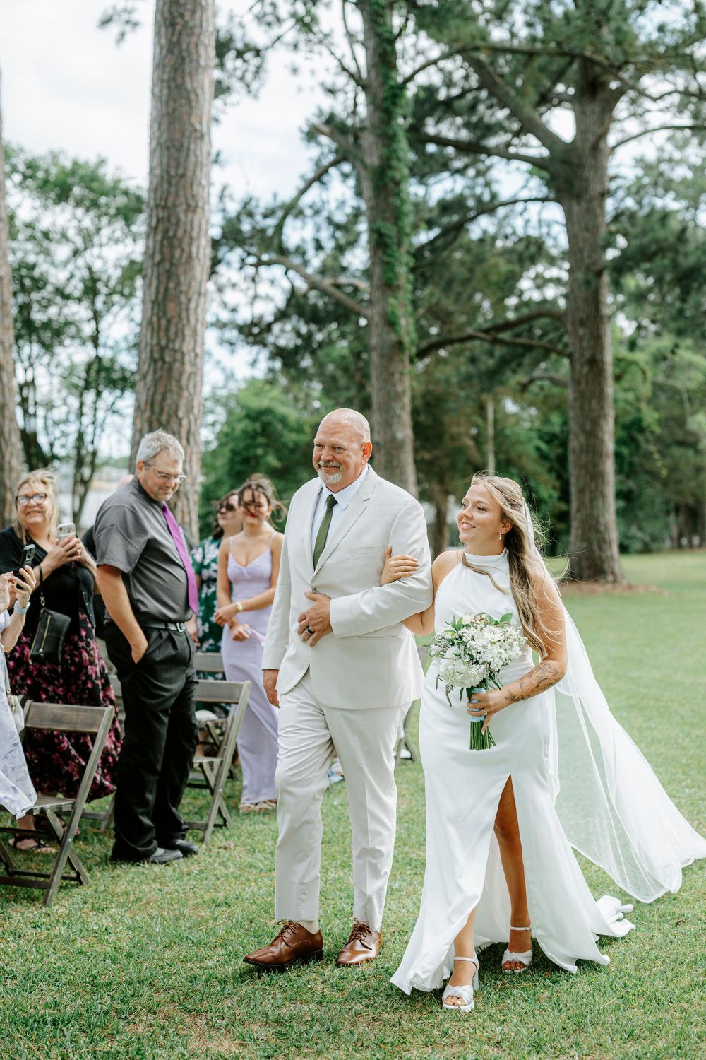 Bride walks down outdoor aisle with escort in white suit, holding bouquet, surrounded by guests seated on chairs.