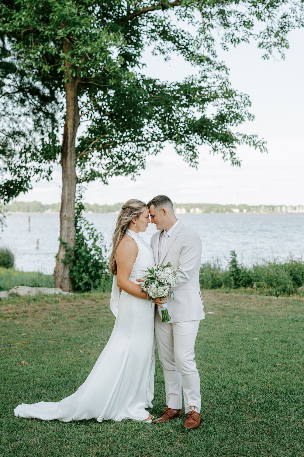 Bride and groom share a romantic forehead touch near a waterfront, she in white gown holding bouquet, he in beige suit.