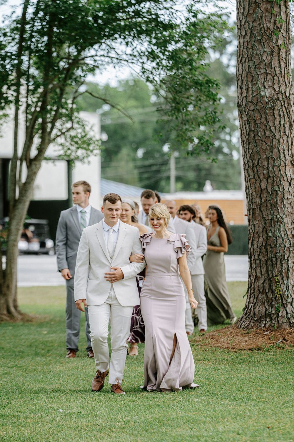 Groom in tan suit escorts bridesmaid in mauve gown as wedding party walks across grass under trees.