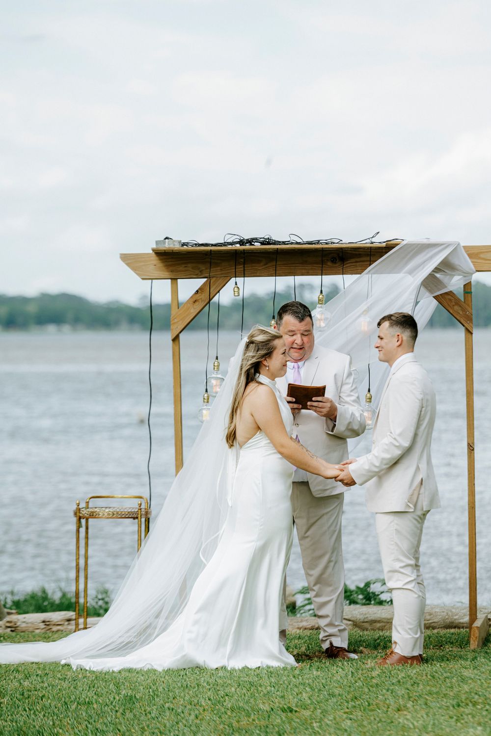 Bride and groom exchange vows under wooden arch with draped fabric by waterfront with officiant.
