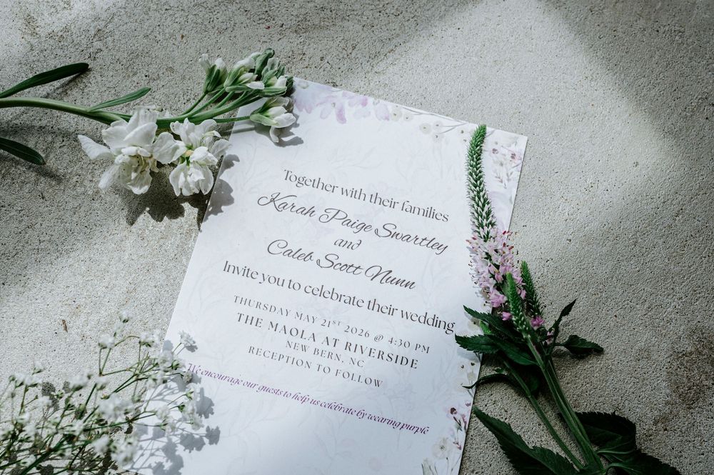 Wedding invitation with floral accents laid flat on surface surrounded by white flowers.