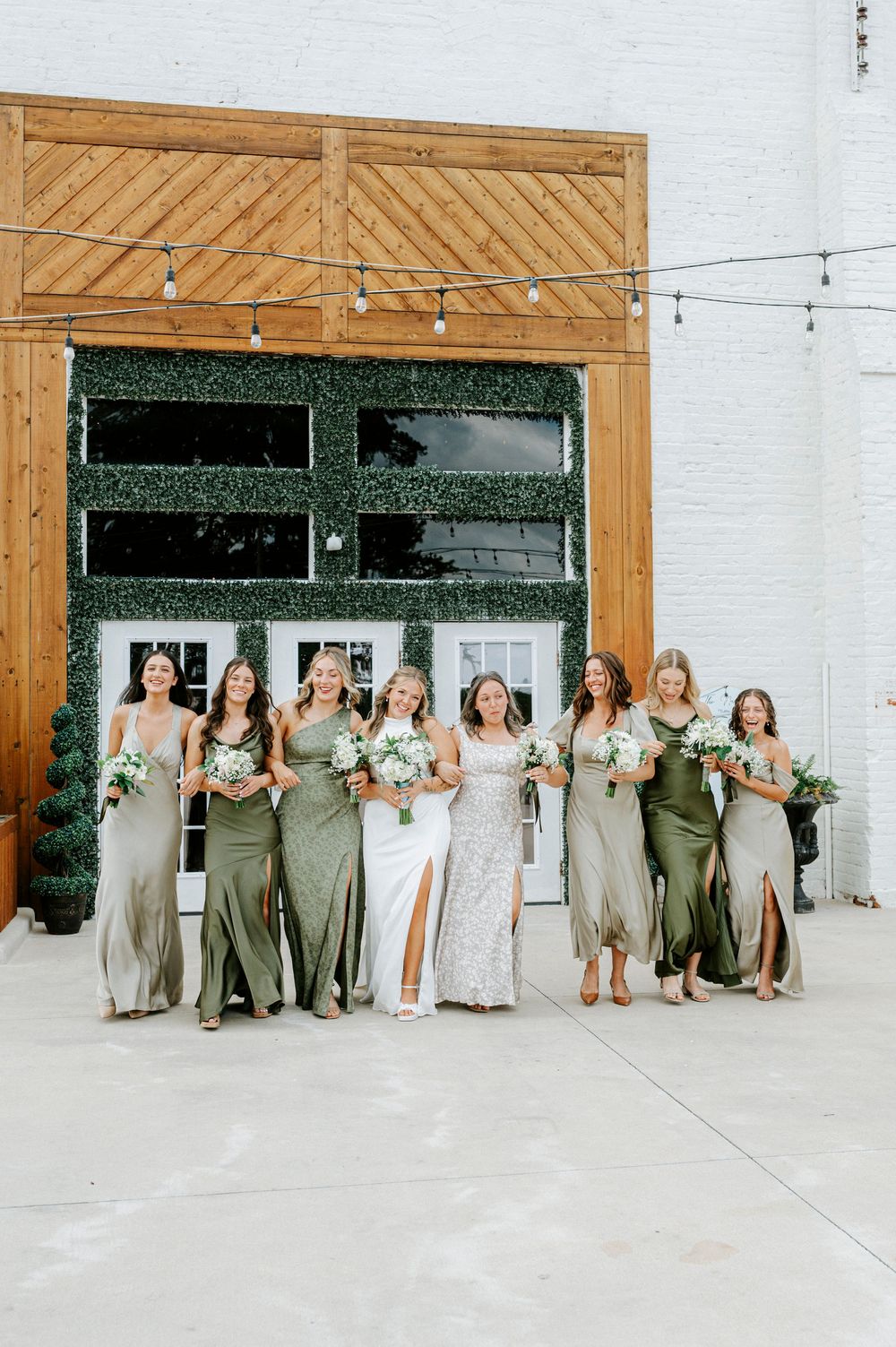 Bride in white lace gown poses with seven bridesmaids in sage green dresses outside rustic venue.
