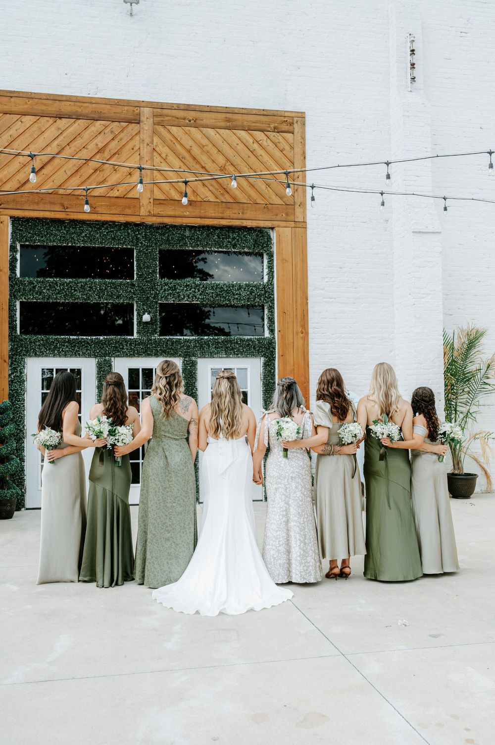 Bride stands with bridesmaids in mixed sage green dresses, backs to camera, outside rustic venue.