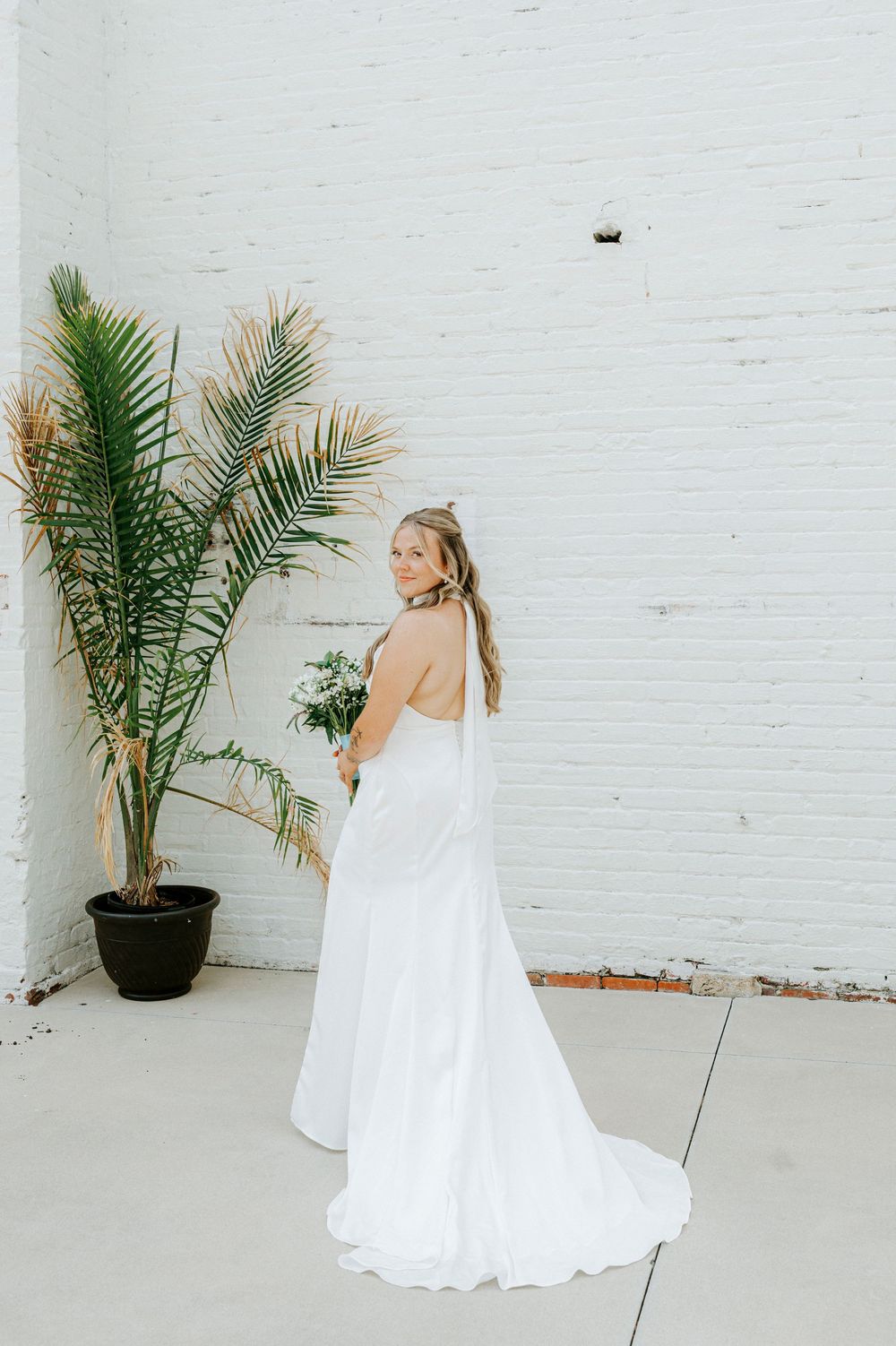 Bride in elegant white backless gown with train poses beside tropical palm plant against white brick wall.