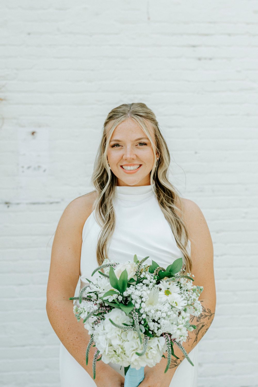 Smiling bride in sleeveless white gown holds white hydrangea and baby's breath bouquet against brick wall.