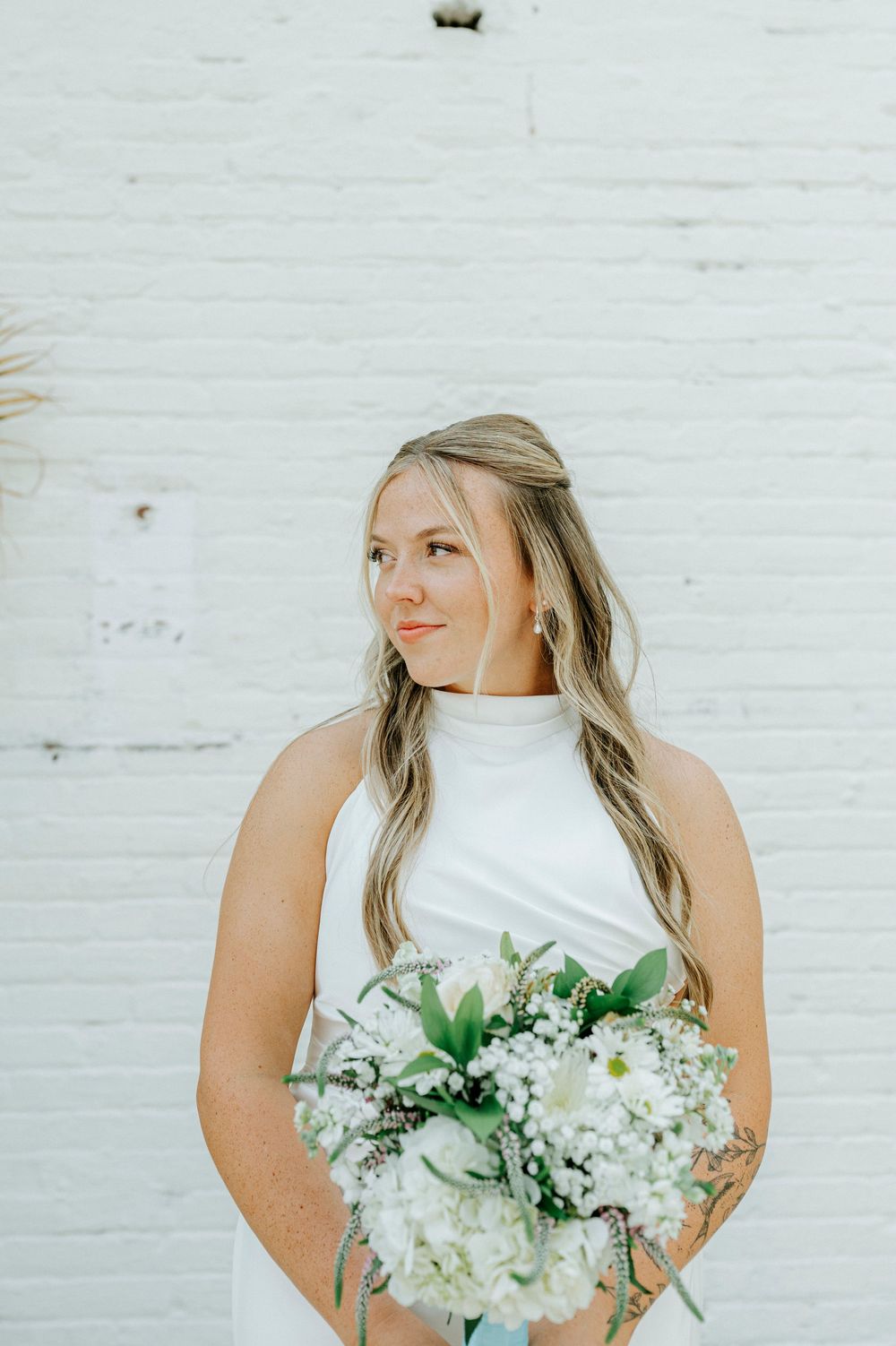 Bride in white halter gown holds white floral bouquet with greenery against a white brick wall.