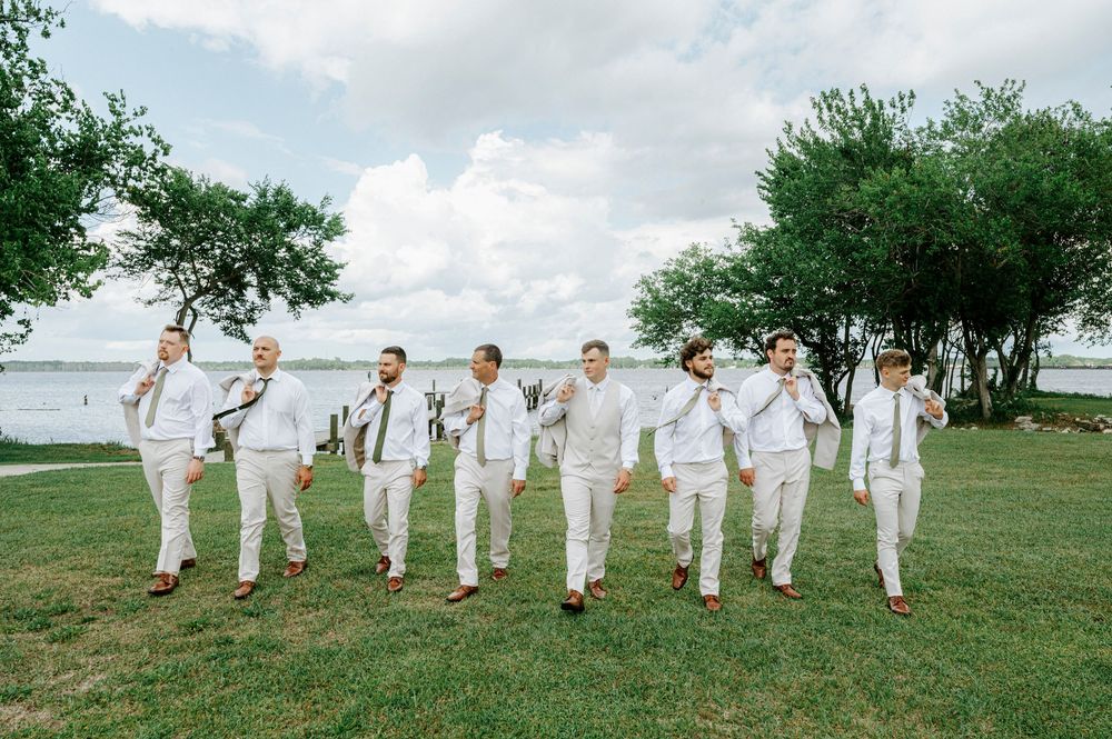Groom and groomsmen in white suits walk together on green lawn near waterfront on wedding day.