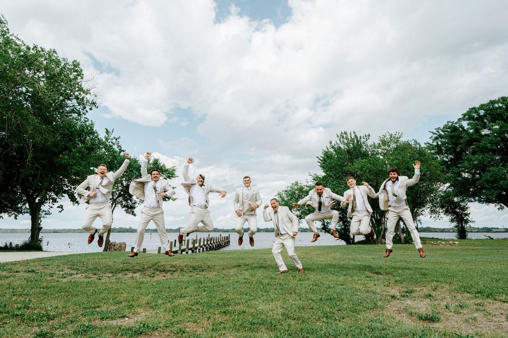 Groomsmen and groom joyfully jumping in air together on green lawn with waterfront backdrop.