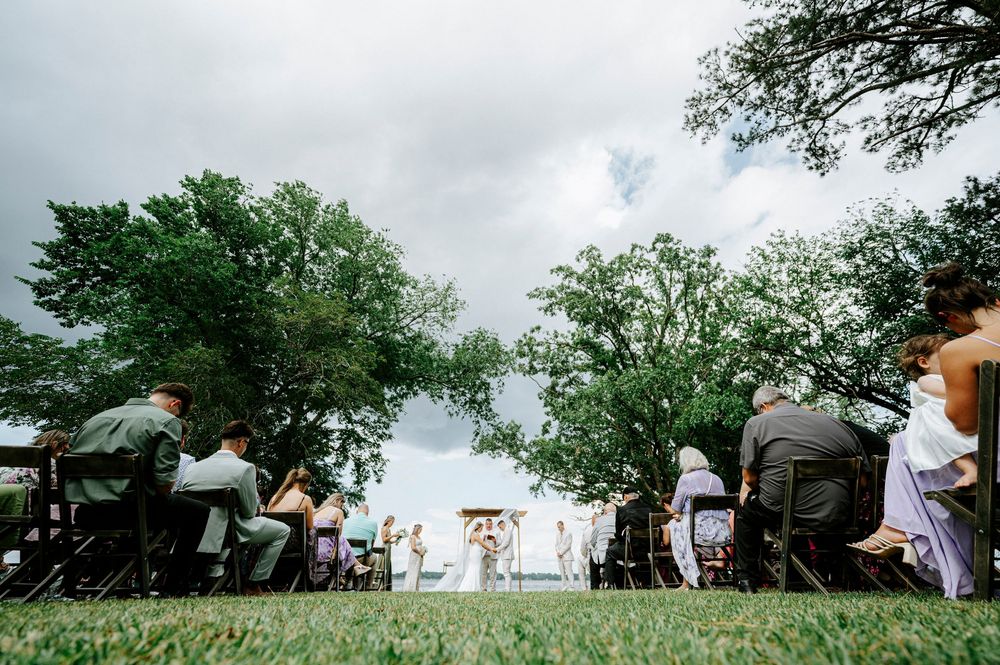 Wide shot of outdoor wedding ceremony with guests seated on chairs under cloudy sky near trees.