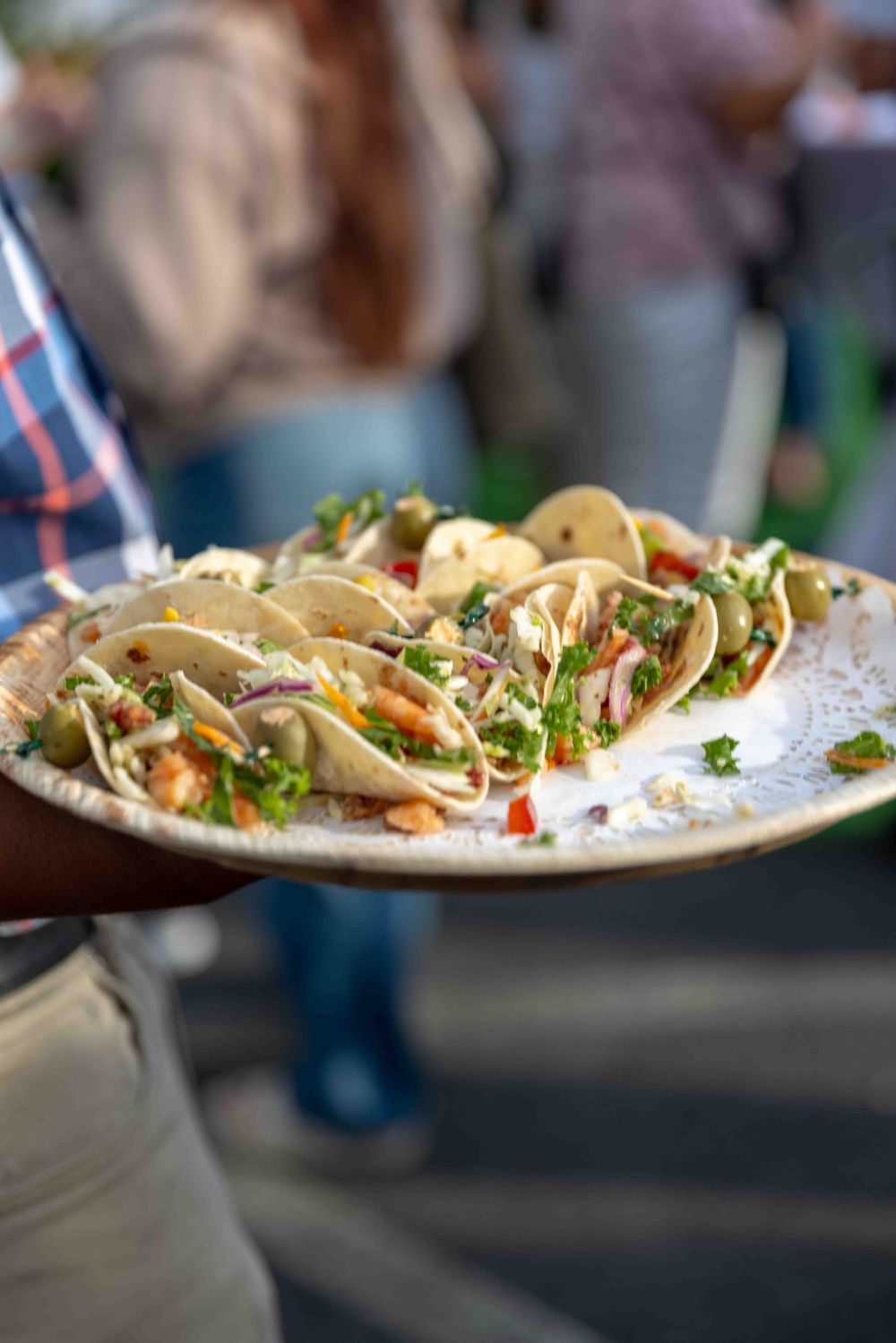 A person holding a plate of fresh street tacos topped with cilantro, red onion, and colorful vegetables at an outdoor event.