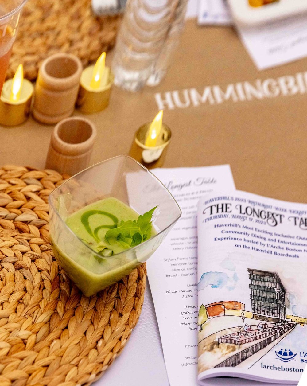 Green gazpacho soup in a clear glass cup at a Hummingbird Longest Table dining event with menu card.