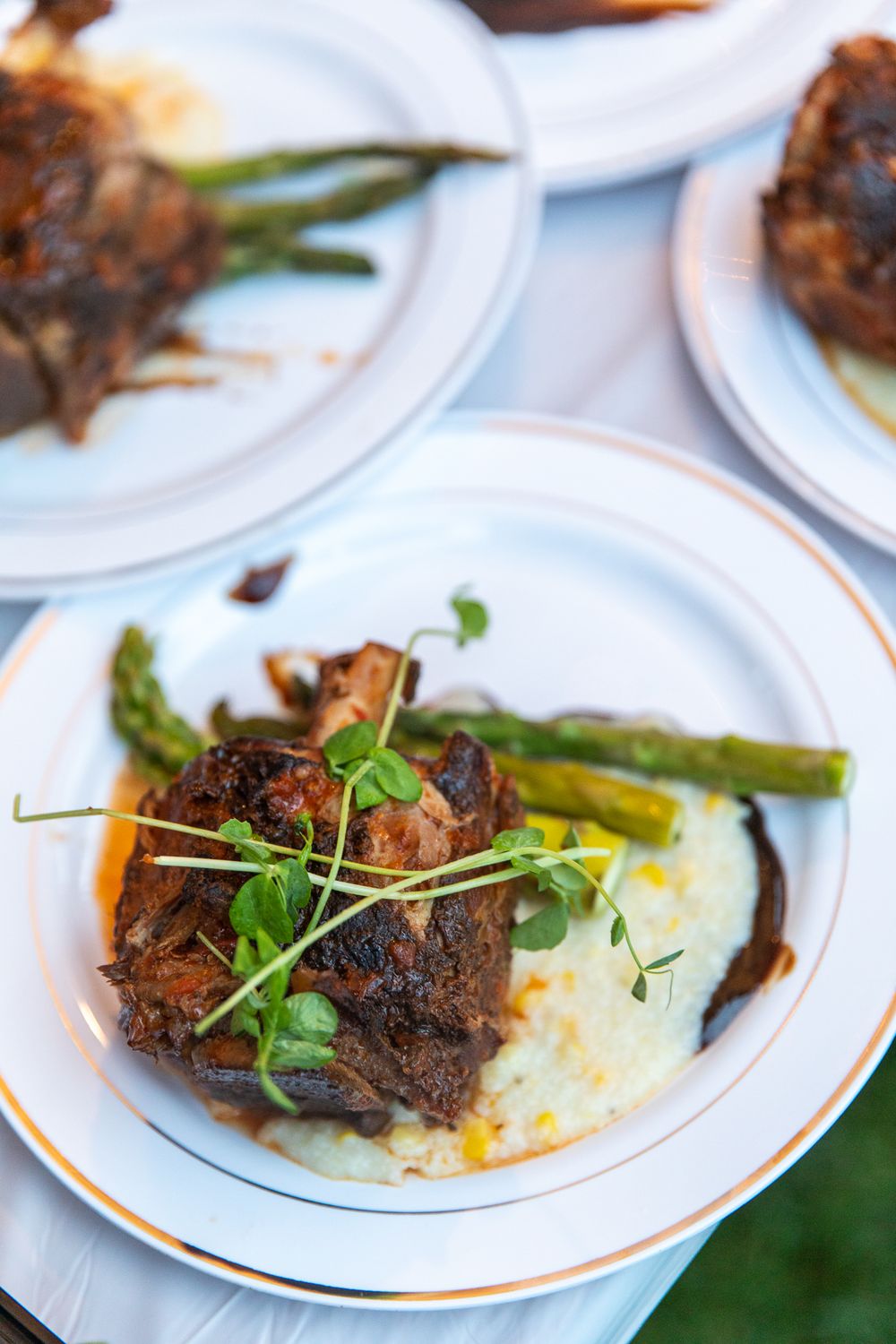 Elegant plated dish with braised meat, creamy grits, roasted asparagus, and fresh microgreens on a white plate.