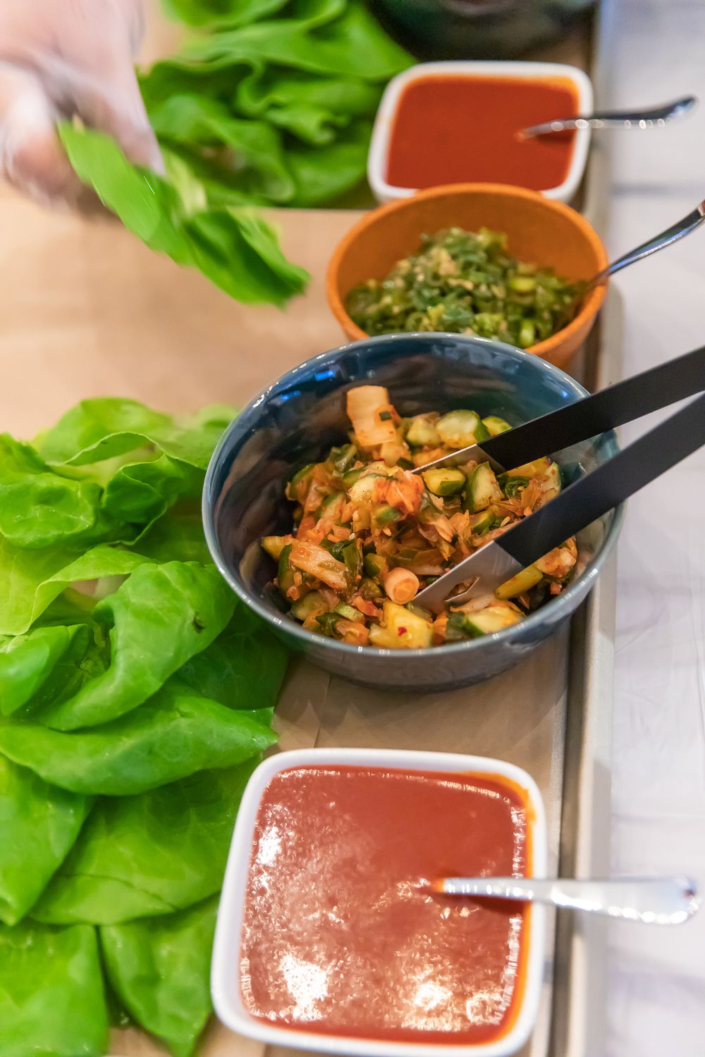 Fresh lettuce wraps with kimchi, dipping sauces, and tongs on a wooden board at a Korean restaurant.