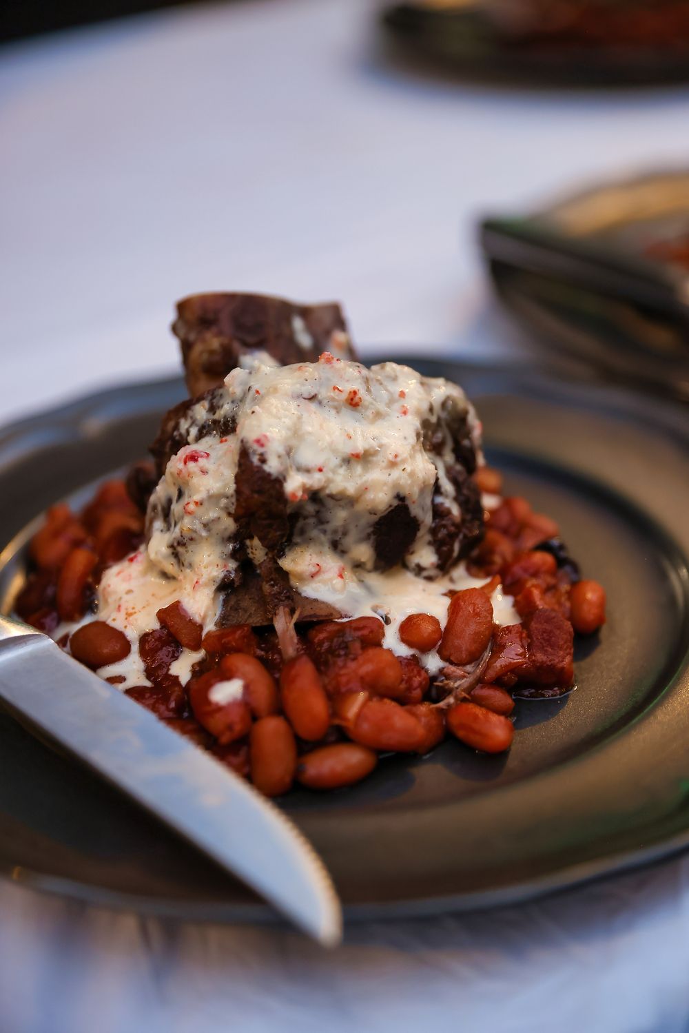 Braised beef short rib served over red beans topped with creamy white sauce on a dark ceramic plate.