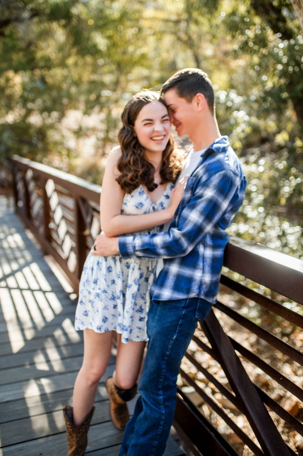 Laughing couple embraces on a wooden bridge surrounded by lush autumn trees in warm golden sunlight.