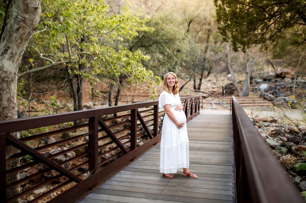 Pregnant woman in white maternity dress standing on wooden bridge surrounded by lush green trees.