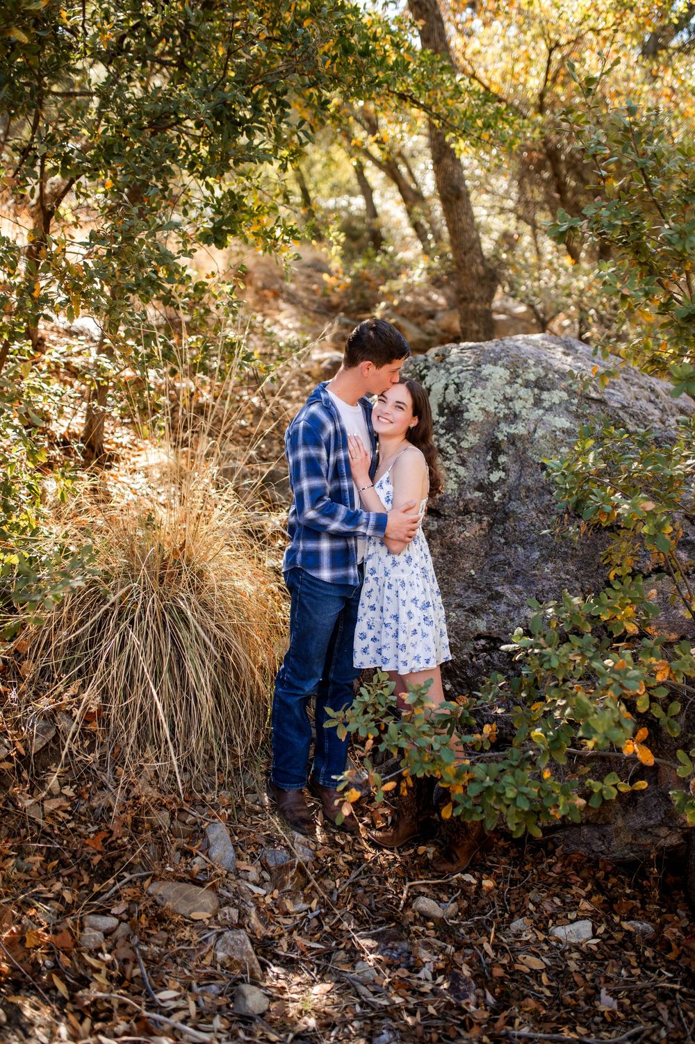Couple shares a tender embrace among autumn foliage and large boulders in a sun-dappled woodland setting.