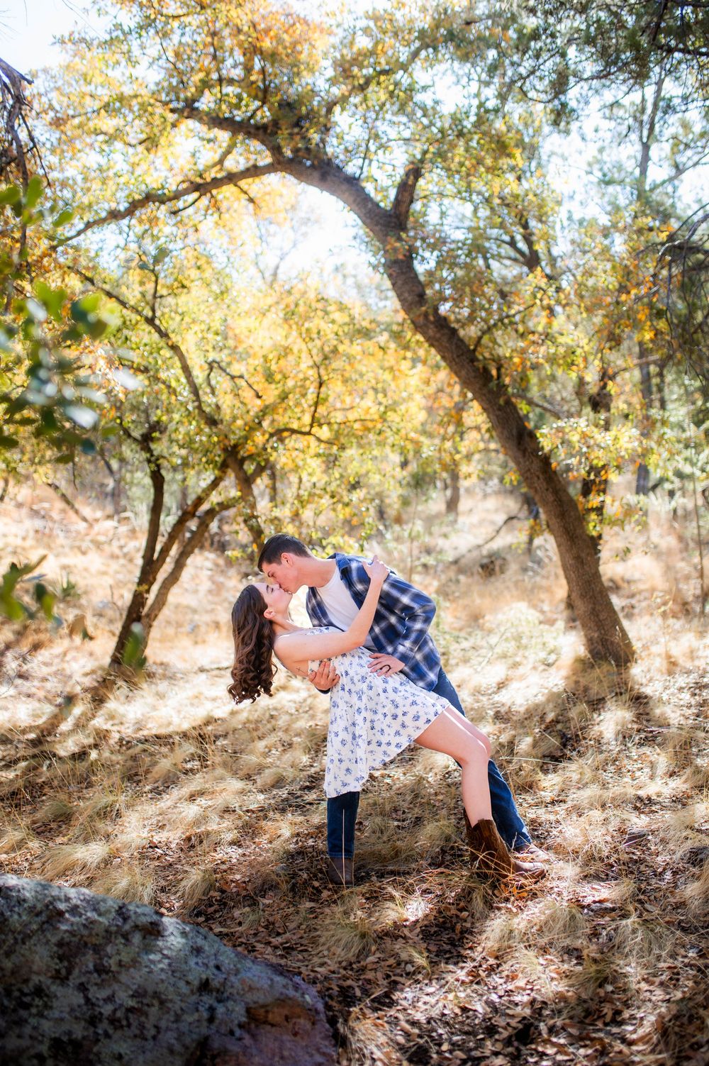 Couple shares a romantic dip kiss in a sunlit autumn forest with golden trees and rocky terrain.