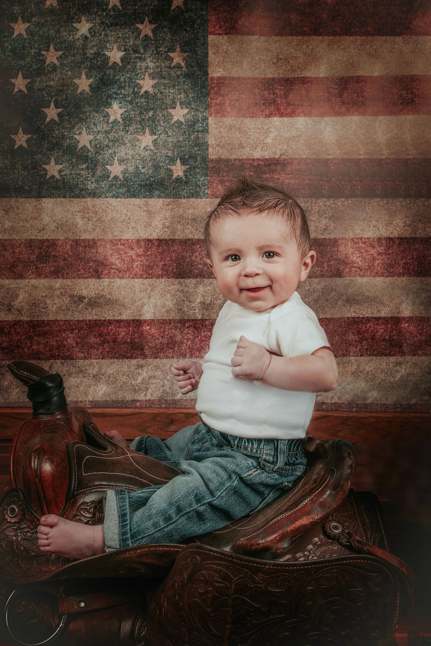 Smiling baby in white onesie and jeans sitting on a Western saddle in front of a vintage American flag backdrop.