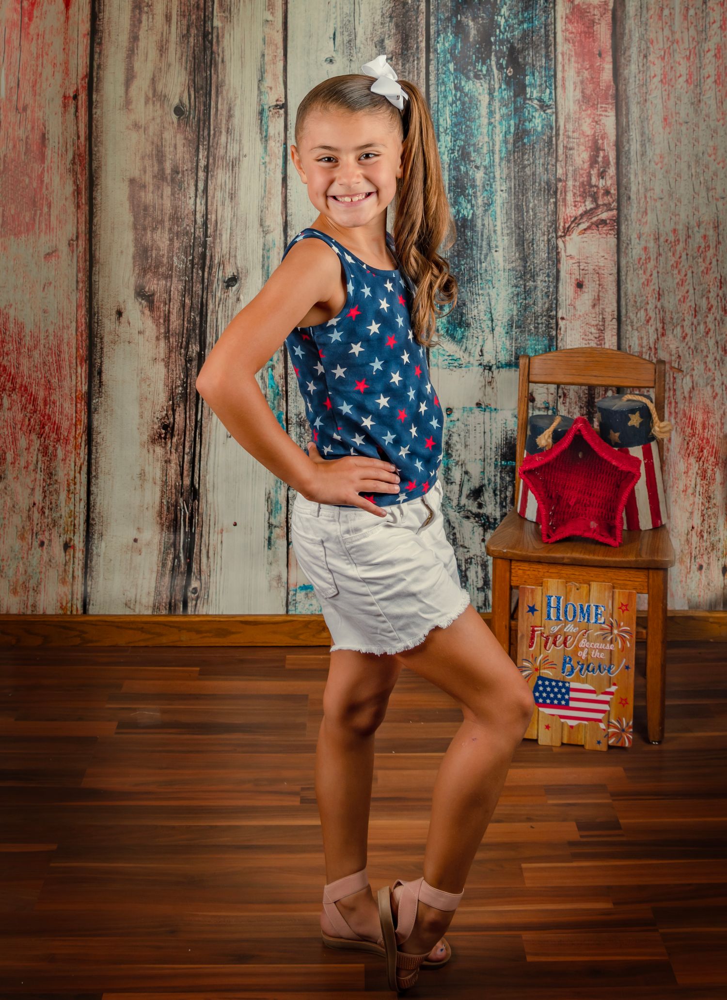 Young girl in patriotic star-print top and white shorts poses confidently against rustic wooden backdrop with American decor.